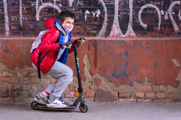 Boy in red jacket wearing a mask riding a scooter by a wall covered in graffiti, illustrating moments karma came instantly.
