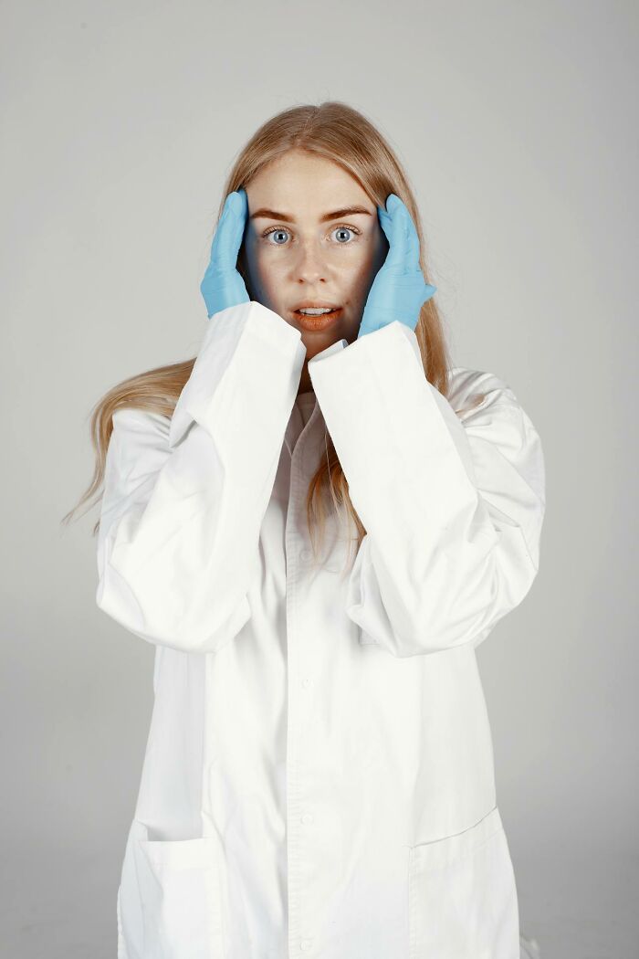 Young woman in a white lab coat and blue gloves showing an embarrassed facial expression indoors on a gray background