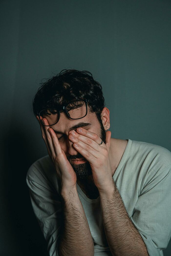 Man covering face with hands, wearing glasses and a grey shirt, expressing stress in a dimly lit environment, true crime fan.