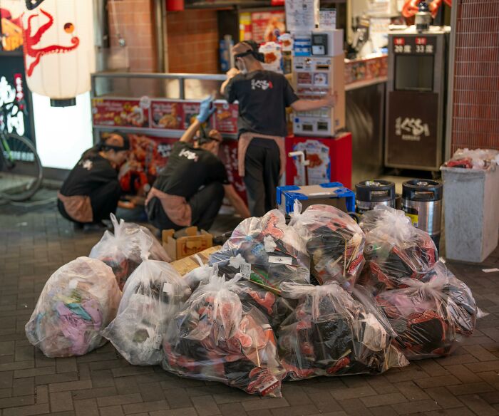 Garbage bags piled outside a restaurant while workers in uniform attend to tasks, illustrating workplace rules annoyance.