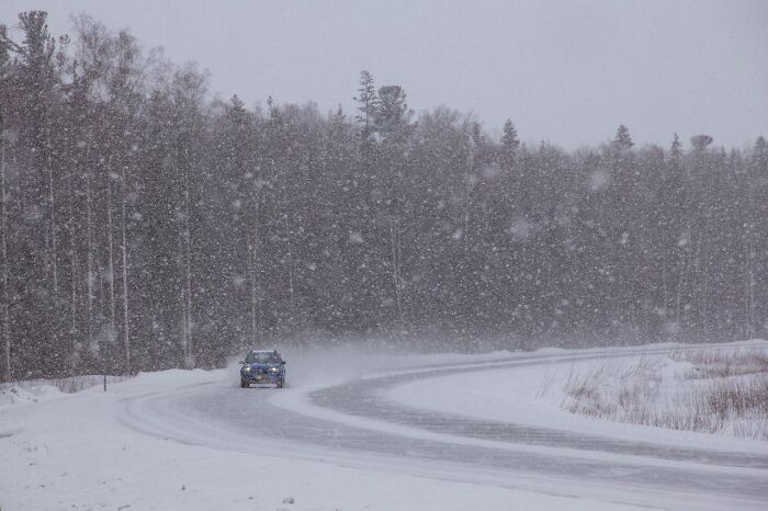 Coche conduciendo por carretera nevada en bosque durante tormenta de nieve, reflejando momentos de karma instantáneo.