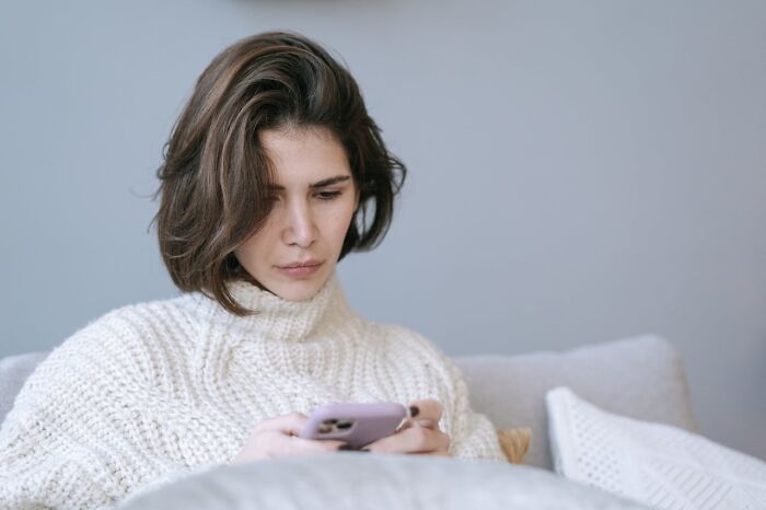 Young woman in a white sweater looking upset while holding a smartphone, reflecting trust broken by close friends.