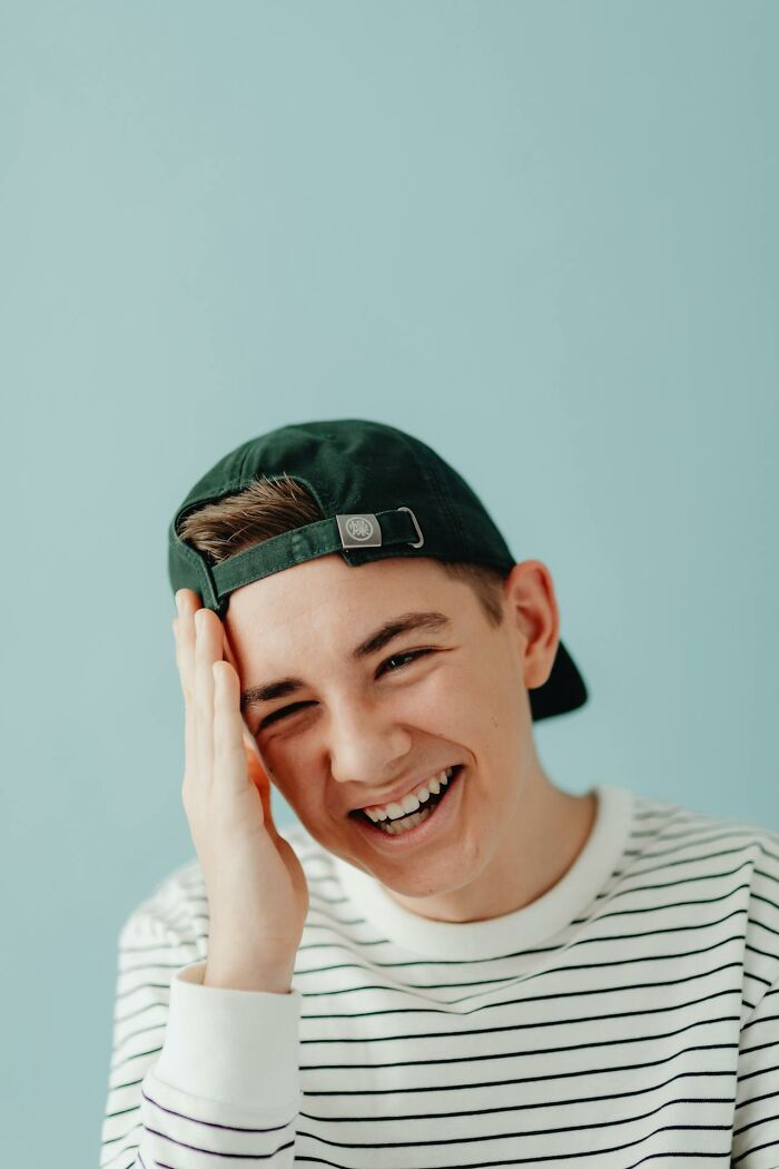 Young man in a striped shirt and cap laughing, expressing joyous reaction to stories of karma striking with zero mercy.