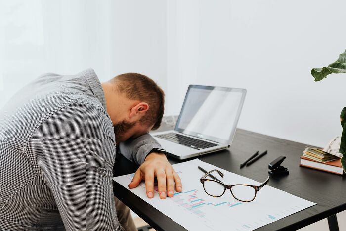 Man resting head on desk in frustration, surrounded by laptop and documents, illustrating annoying workplace rules.