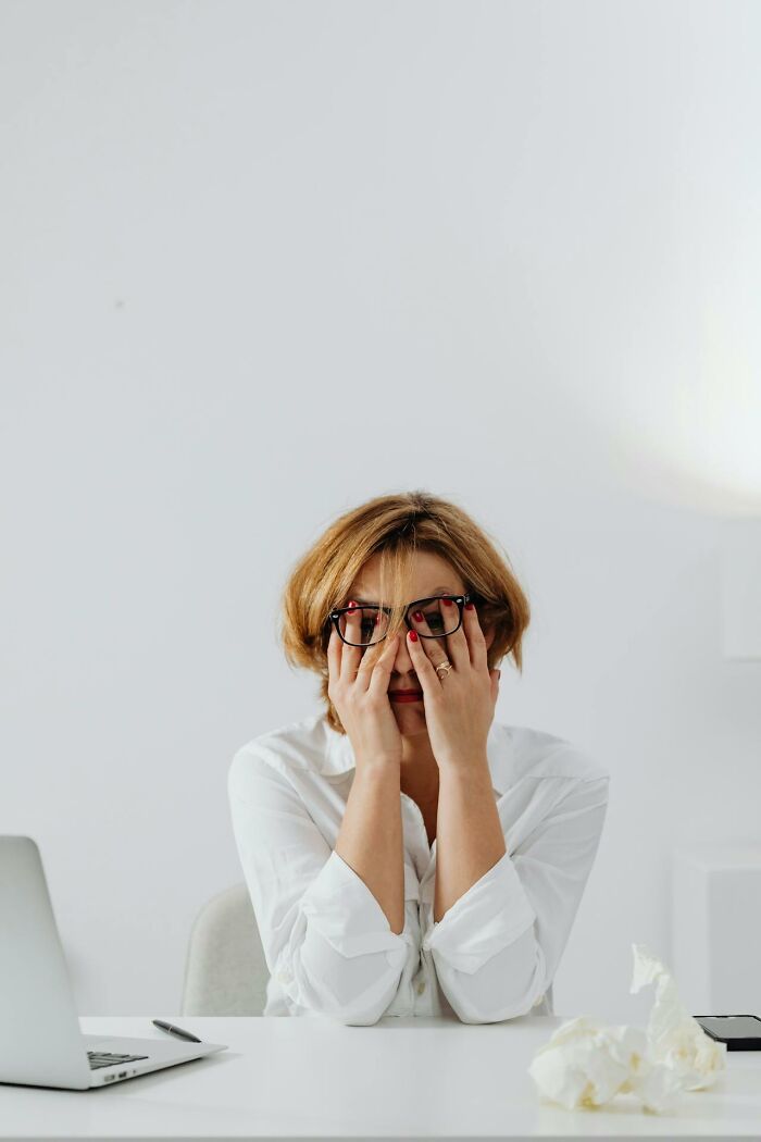 Woman wearing glasses and white shirt sitting at desk stressed with hands on face, illustrating subtle sexism issues for men.