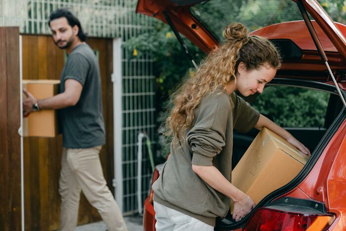 A young woman loading a cardboard box into a car trunk while a man carries another box outside, moving boxes with care.