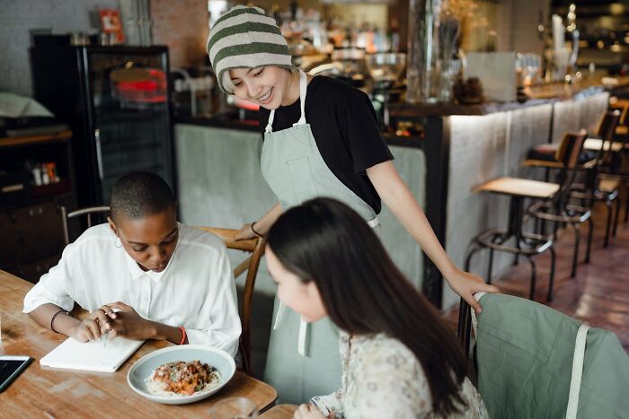 Three people at a café with one person smiling while serving food, capturing a casual moment of embarrassment and laughter.