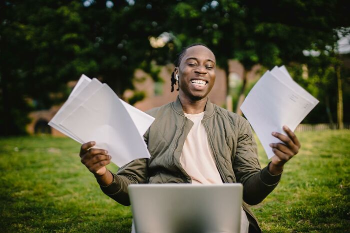 Young man smiling outdoors, holding papers and working on a laptop, illustrating painful table for two moments shared by waiters.