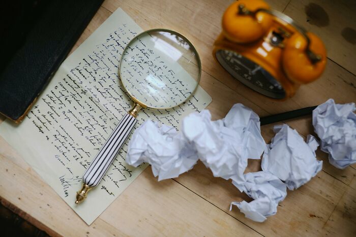 Magnifying glass over handwritten letter with crumpled papers and retro alarm clock on wooden table, recognizing characters.