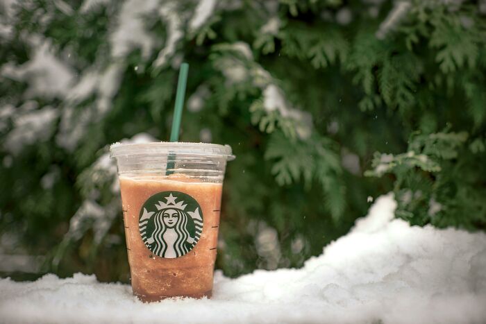 Iced Starbucks coffee cup with green straw placed on snow in front of green foliage, illustrating brand description concept.