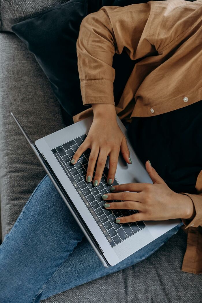 Person using a laptop on a couch, typing with green nails and wearing casual brown shirt and blue jeans, true crime fan concept.