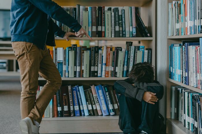 Person pointing at another sitting on the floor with arms crossed, illustrating karma striking in a library setting.
