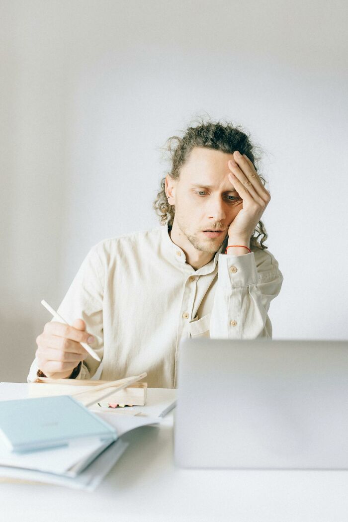 Young man looking stressed while using a laptop, surrounded by notebooks and a pen, experiencing karma striking moment.