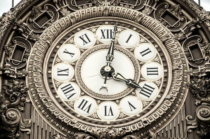 Ornate antique clock face with Roman numerals surrounded by intricate decorative carvings on a historical building facade.
