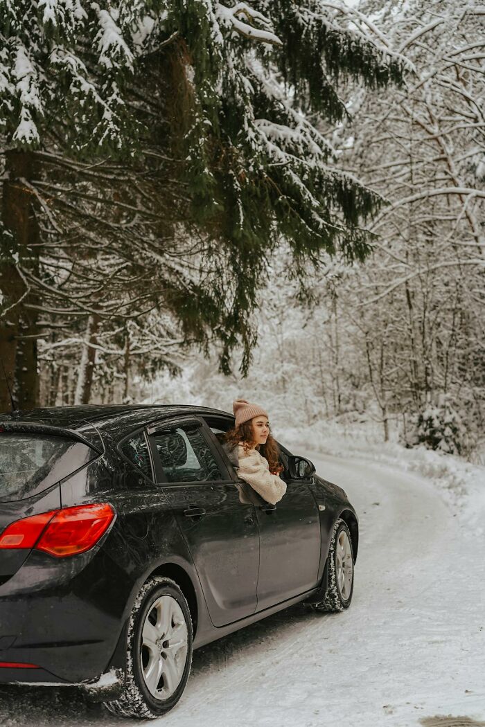 Young woman leaning out of a black car window on a snowy forest road, capturing a serene karma moment in winter.