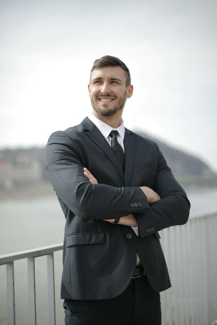 Confident man in suit standing outdoors near railing smiling with arms crossed, representing karma striking with zero mercy.