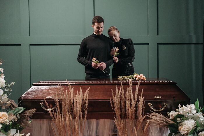 Man and woman mourning at a funeral standing by a closed casket holding flowers, illustrating funeral stories and emotions.