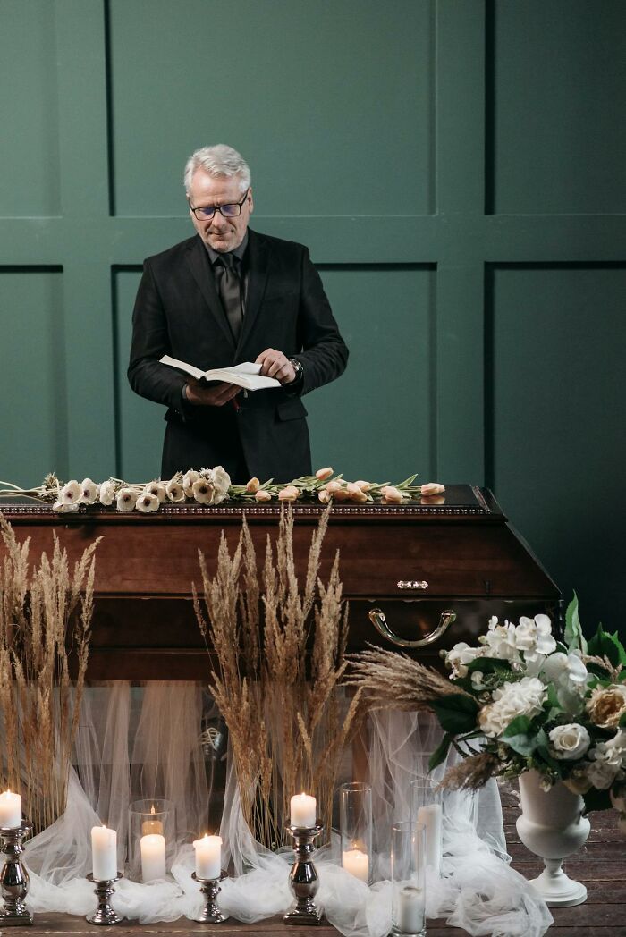 Man in black suit reading from a book beside a coffin with flowers and candles, funeral stories setting.
