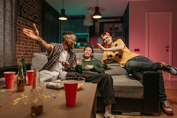 Three friends hanging out in a living room, sharing chaotic and dramatic breakup stories while enjoying drinks.