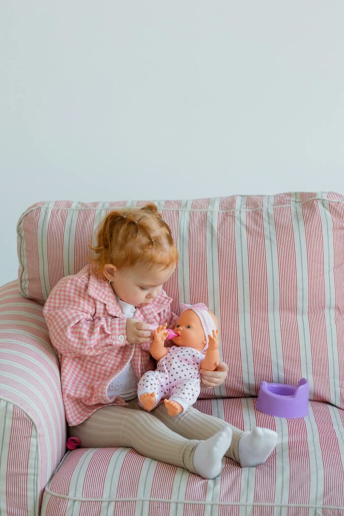 Toddler girl playing with baby doll on striped couch, illustrating subtle sexist stereotypes in childhood behavior.