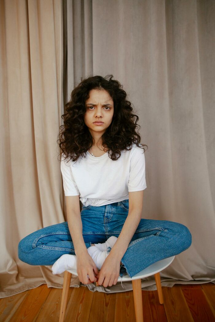 Young woman with curly hair and serious expression sitting cross-legged on a chair, reflecting trust broken by close friends.