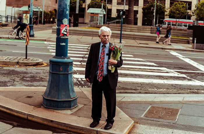 Older man in a suit holding a bouquet of flowers, standing alone on a city street corner near a crosswalk.