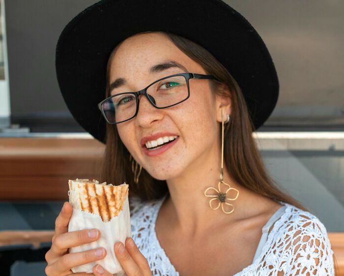 Young woman wearing glasses and a black hat eating a wrap, representing restaurant staff stories about the bill and money.