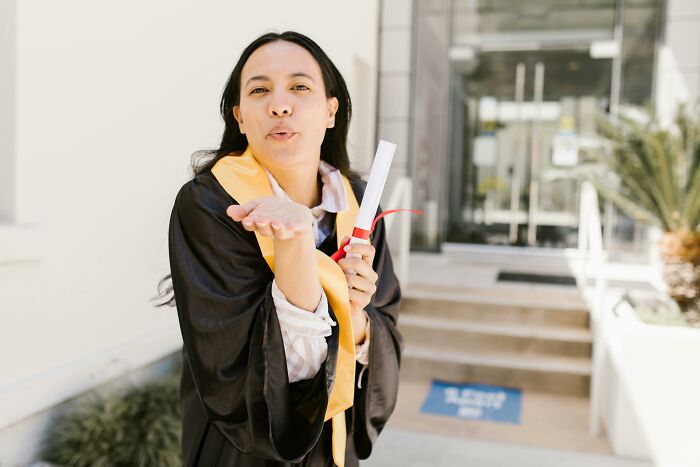 Young woman in graduation gown holding diploma and blowing a kiss outdoors, symbolizing shocking and dark secrets revealed.