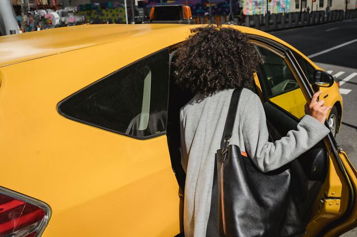 Person with curly hair and a black bag getting into a yellow taxi, capturing moments of embarrassing and hilarious stories.