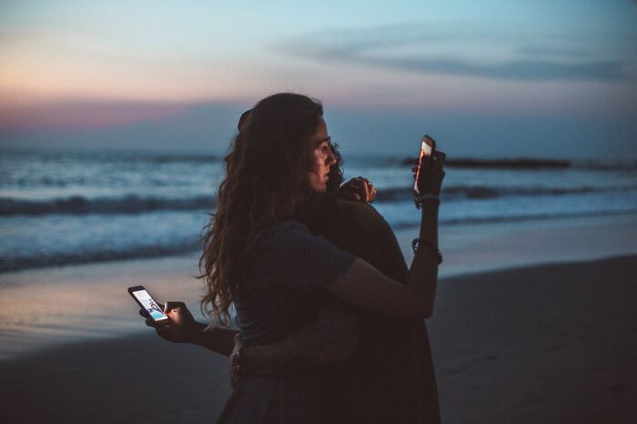 Couple at the beach at dusk, both secretly looking at their phones, illustrating first signs someone is having an affair.