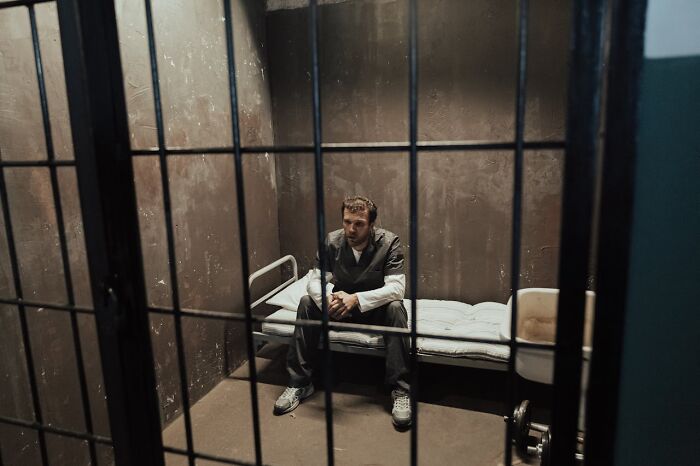 Man in prison cell wearing gray uniform sitting on bed with hands clasped, reflecting true crime fan intrigue.