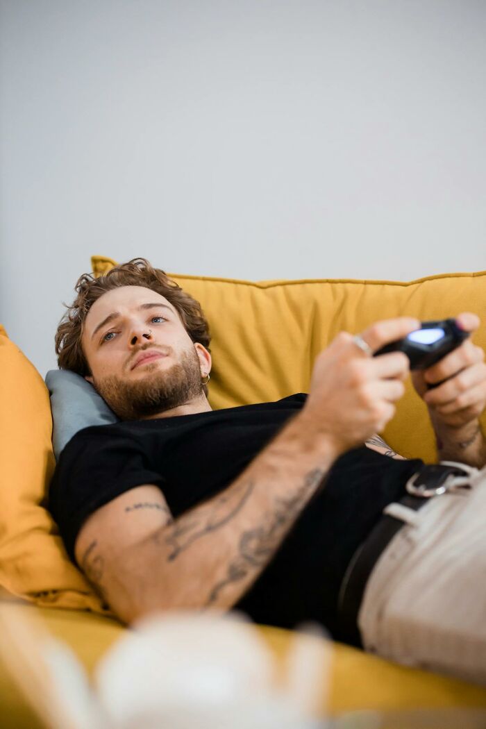 Young man with tattoos lying on yellow couch, holding a game controller, representing secrets people hide from partners.