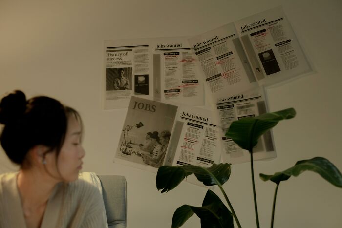 Woman looking at wall covered with job listings and newspaper clippings, hinting at shocking and dark secrets affecting lives.