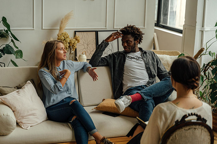 Couple having a tense conversation on a couch during a therapy session with a female therapist taking notes nearby.