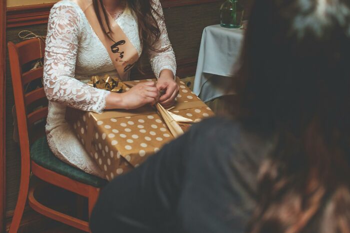 Woman in a white dress sitting at a table with a large gift box, depicting a painful table for two moment ending alone.