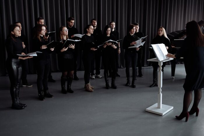 Choir dressed in black rehearsing somber funeral songs under conductor's direction in a dimly lit room.