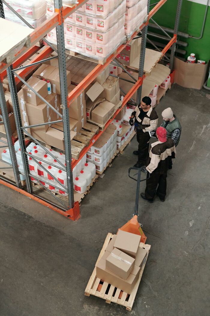 Three workers in a warehouse organizing boxes and containers on shelving, illustrating teamwork and karma concepts.