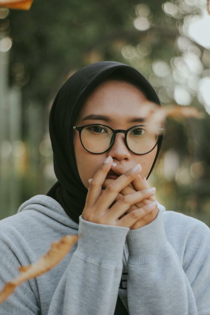 Young woman wearing glasses and a black hijab, covering her mouth with hands, reflecting on worst and craziest funeral stories.