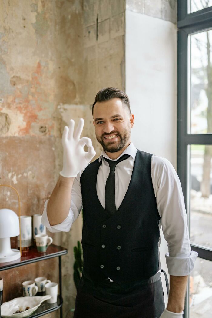 Man in black vest and white gloves smiling and making okay sign indoors related to people embarrassed moments.