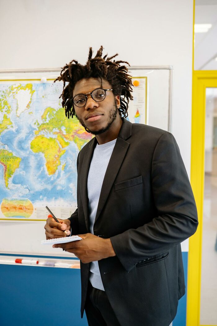 Young man with dreadlocks and glasses writing in a notebook near a world map, capturing hilarious embarrassing moments.