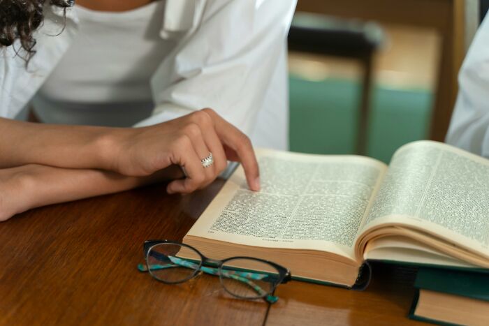 Person pointing at an open dictionary page, studying word origins and testing etymology skills on a wooden table.