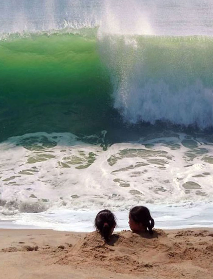 Two kids buried in sand at the beach with a huge wave about to crash behind them, a hilarious photo before disaster struck.