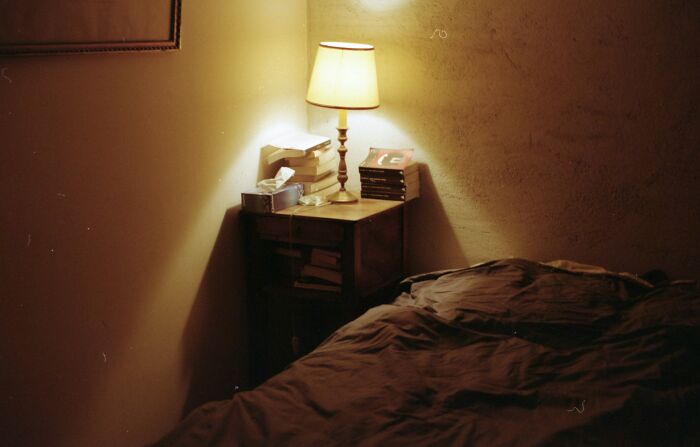 Dimly lit bedroom with a bedside table, lamp, books, and a rumpled bed, evoking memories of disastrous playdates.