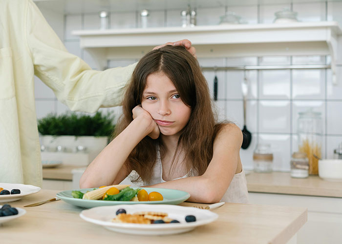 Young girl looking upset at a kitchen table with healthy food while a man gently places his hand on her head.