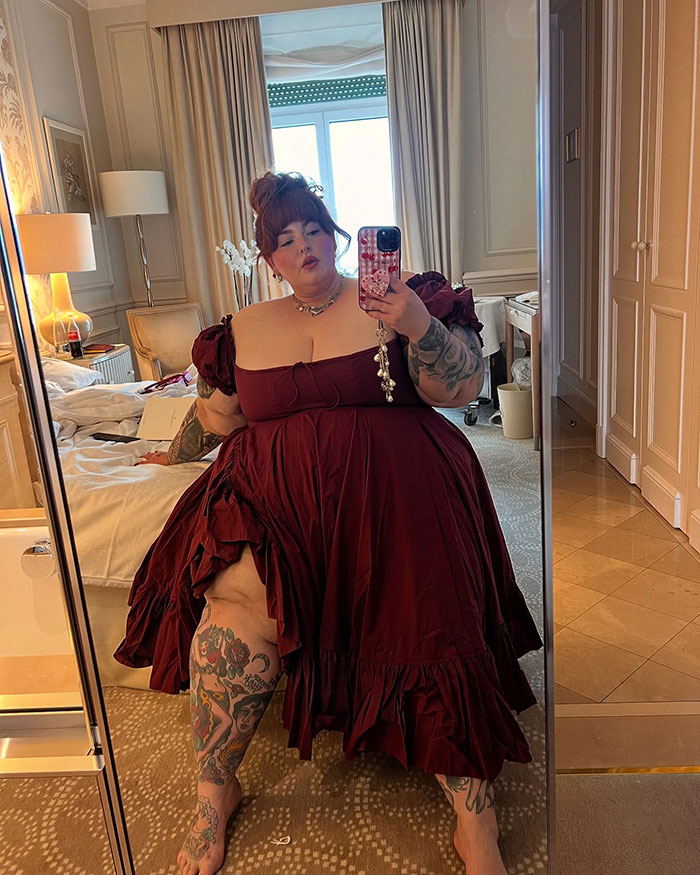 Plus-size model in a burgundy dress taking a mirror selfie, showcasing tattoos and confident pose indoors.