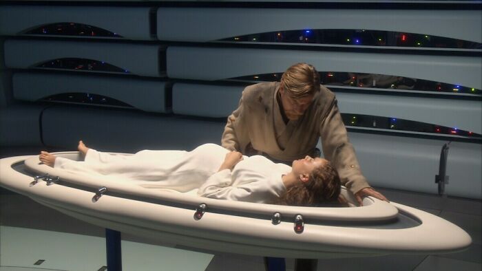 Scene from a movie showing an unrealistic women trope with a woman lying on a futuristic medical bed and a man leaning over her.