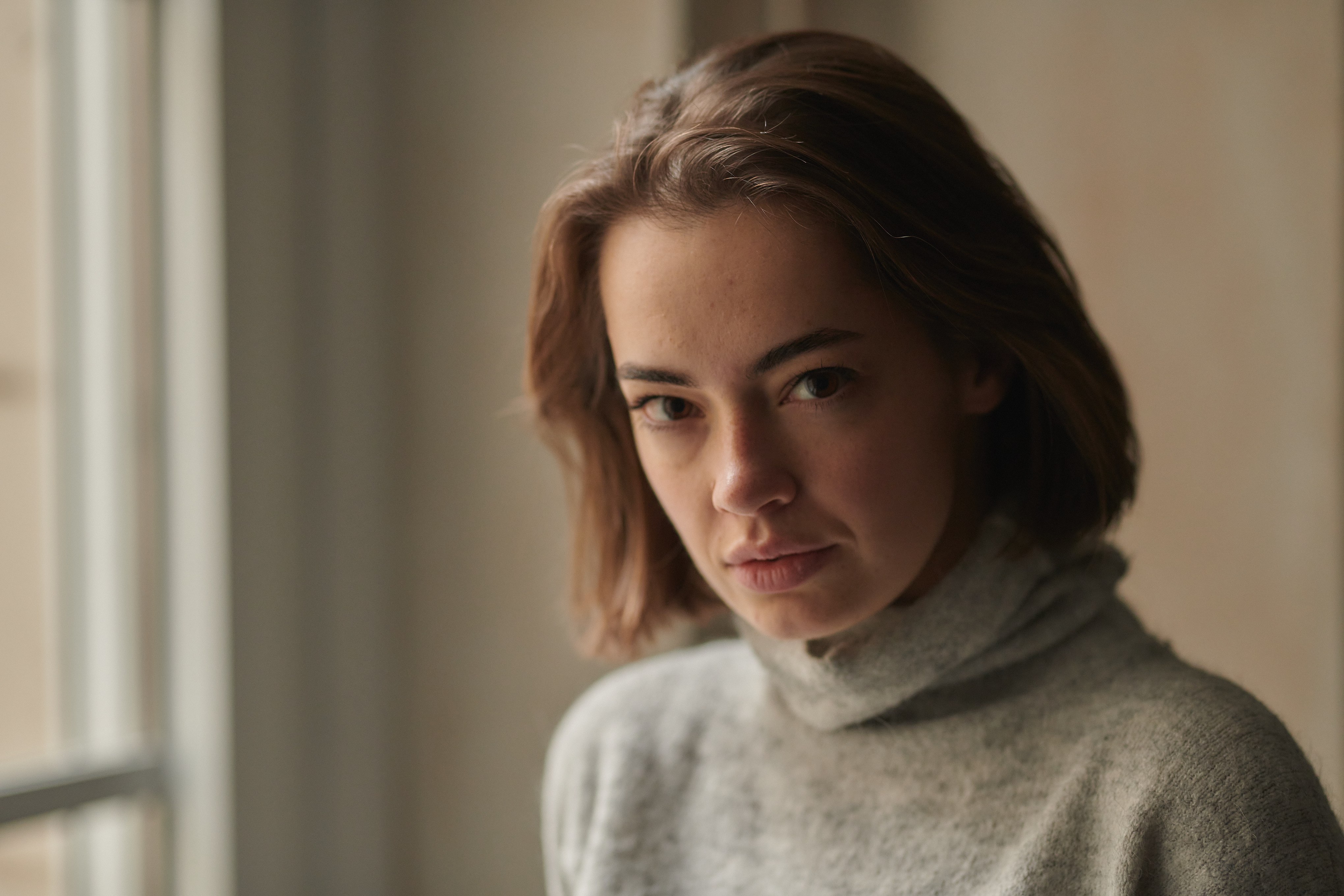 Young woman with short brown hair and gray turtleneck sweater looking serious by a window indoors.