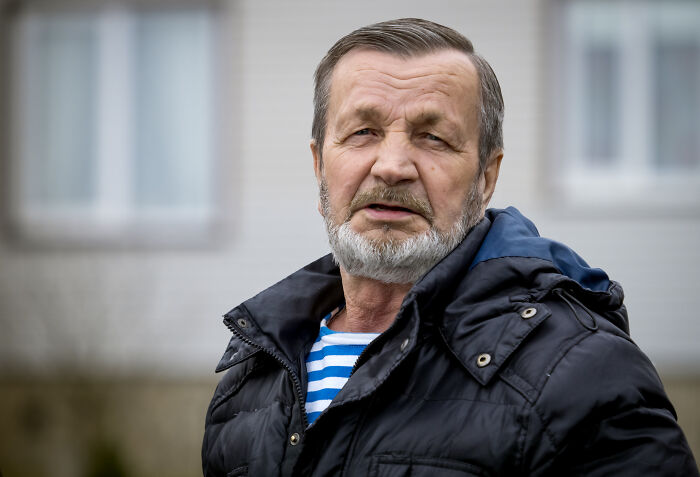 Older man with gray beard wearing a black jacket and striped shirt standing outside during daytime in a residential area