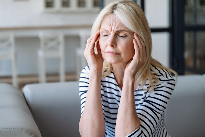 Woman in striped shirt sitting on couch with eyes closed and hands on temples showing stress from worst first opinions doctors heard.