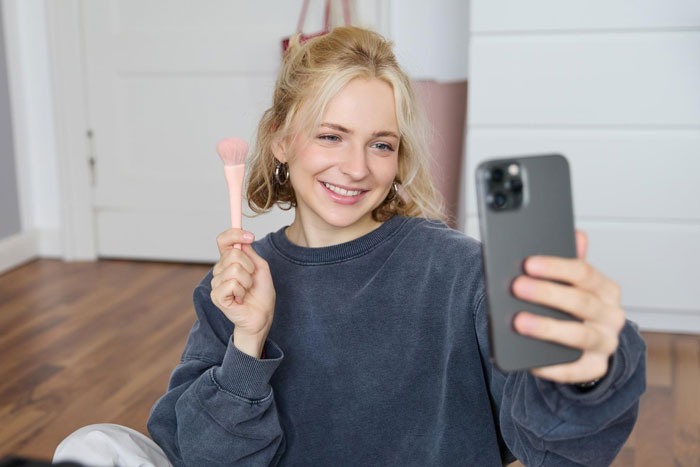 Young woman smiling while taking a selfie with a makeup brush, relating to vegan sister and leather shoes story.